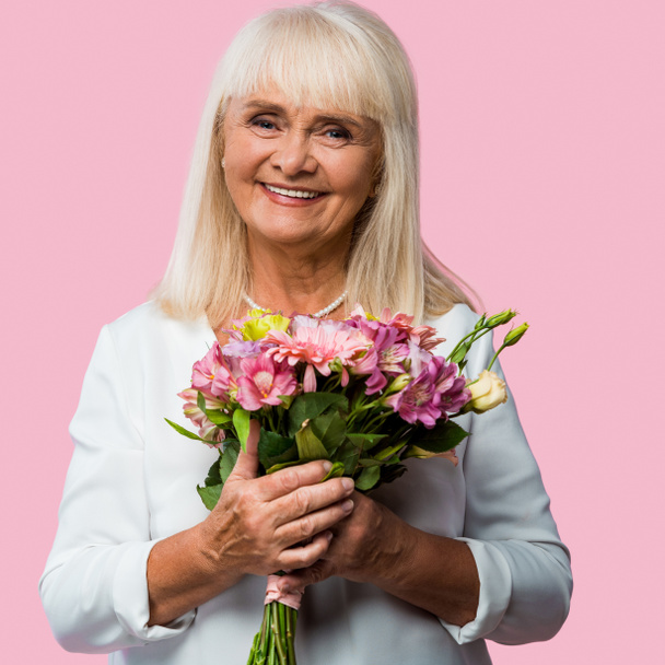 mujer mayor positiva sosteniendo ramo de flores en flor aislado en rosa
  - Foto, Imagen