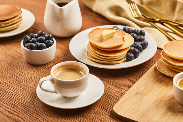 sabroso desayuno con taza de café, panqueques y arándanos cerca de tela de lino con cubiertos en la superficie de madera
 - Foto, Imagen