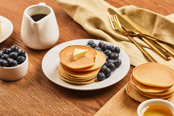 plate with pancakes and blueberries, jug with syrup near linen cloth with golden cutlery on wooden surface  - Photo, Image