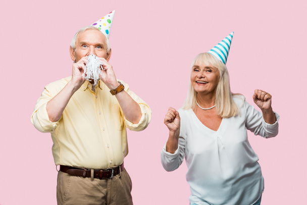 senior man covering face with party blower near cheerful wife in party cap gesturing isolated on pink  - Fotó, kép