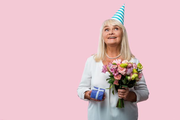 cheerful senior woman in party cap holding flowers and gift box isolated on pink  - Fotó, kép