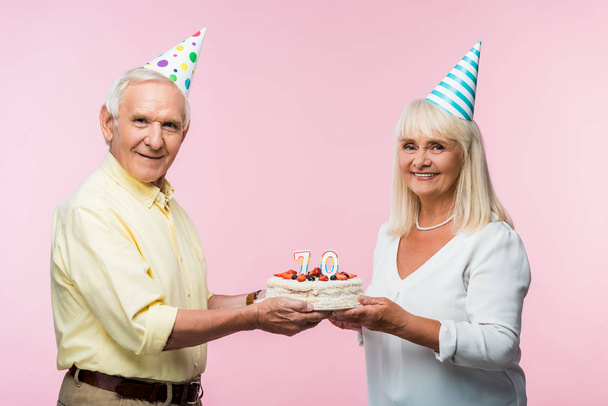 ピンクに隔離されたバースデーケーキを保持するパーティーキャップで幸せなシニアカップル  - 写真・画像