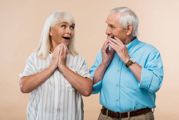verbaasd Senior vrouw kijken naar man bedekken gezicht met handen geïsoleerd op beige  - Foto, afbeelding