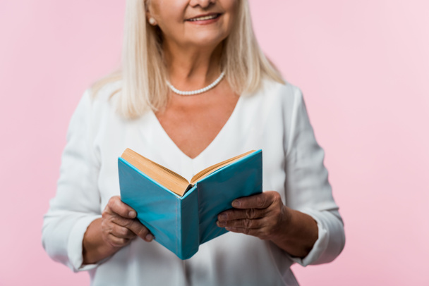 oříznutý pohled na veselou starší ženu držící knihu izolovanou na růžovém  - Fotografie, Obrázek