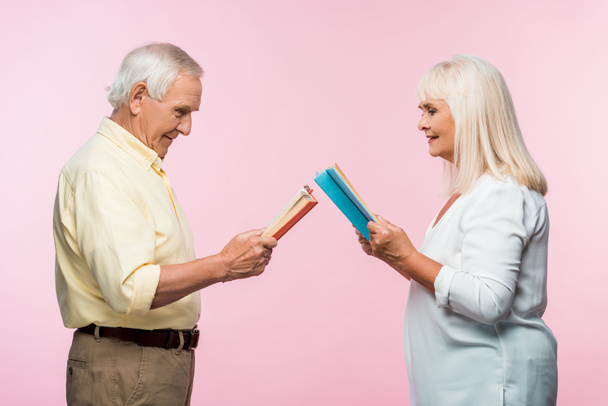 side view of happy senior couple reading books while standing isolated on pink  - Foto, imagen