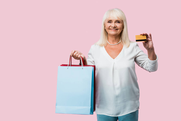 šťastná žena v důchodu s nákupními taškami a kreditní kartou izolovaná na růžovém  - Fotografie, Obrázek