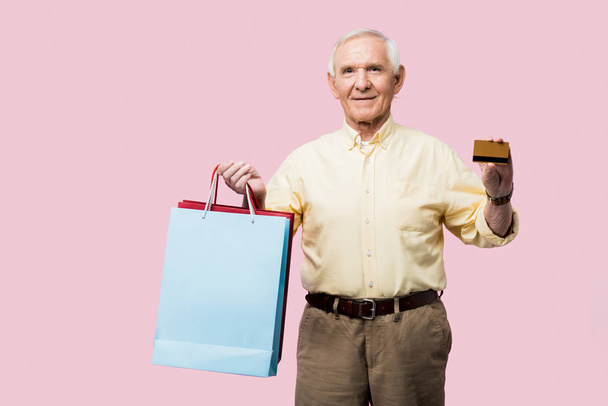 Gelukkig gepensioneerde man houden creditcard en boodschappentassen geïsoleerd op roze  - Foto, afbeelding