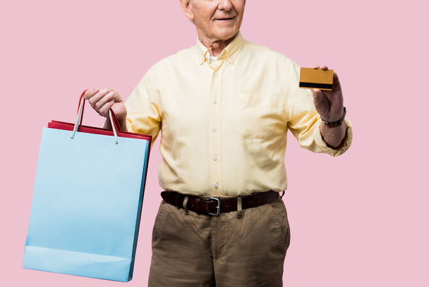 cropped view of happy retired man holding credit card and shopping bags isolated on pink  - Foto, afbeelding