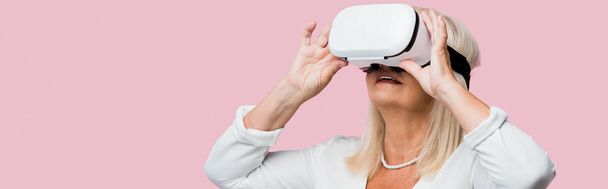 panoramic shot of retired woman touching virtual reality headset isolated on pink  - 写真・画像