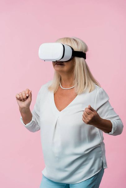 Senior vrouw dragen Virtual Reality headset en gebaren geïsoleerd op roze  - Foto, afbeelding