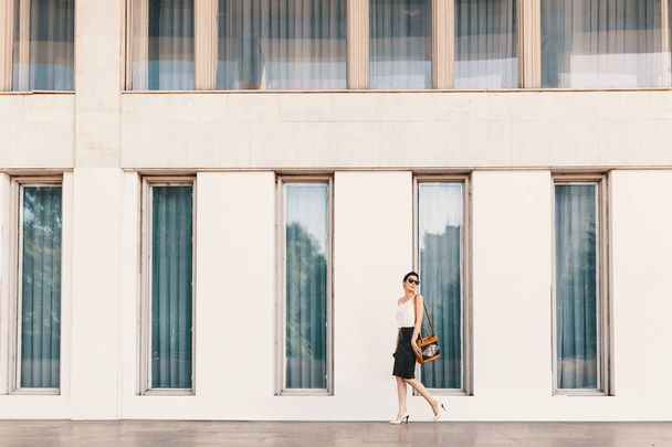 Muodikas pitkä liiketoiminnan nainen aurinkolaseissa kävely lähellä rakennusta, jossa on korkeat ikkunat kaupungissa
 - Valokuva, kuva
