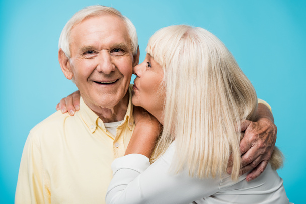 retired woman kissing cheek of cheerful man with grey hair on blue  - Fotoğraf, Görsel