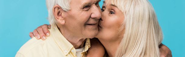 panoramic shot of retired woman kissing cheek of cheerful man with grey hair isolated on blue  - Photo, Image