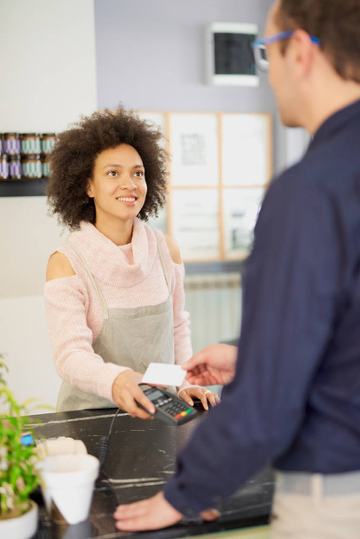 Hymyilevä sekarotuinen nainen leivonnaiskaupan työntekijä veloittaa laskun, kun taas valkoihoinen mies, jolla on luottokortti
. - Valokuva, kuva