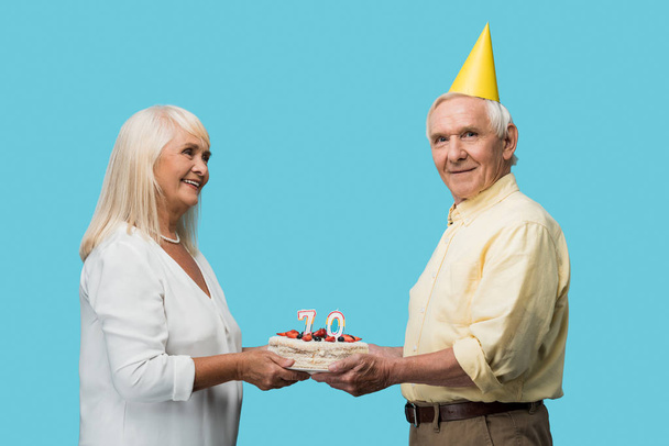 Fröhlicher Rentner mit Partymütze hält Geburtstagstorte neben Ehefrau auf blauem Grund  - Foto, Bild