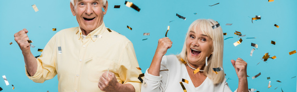 Panoramaaufnahme eines glücklichen Rentnerehepaares, das gestikulierend neben fallendem Konfetti auf blauem Grund steht  - Foto, Bild