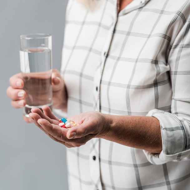 enfoque selectivo de la mujer mayor sosteniendo píldoras y vaso de agua aislado en gris
  - Foto, Imagen