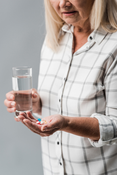cropped view of senior woman holding pills and glass of water isolated on grey  - Foto, immagini