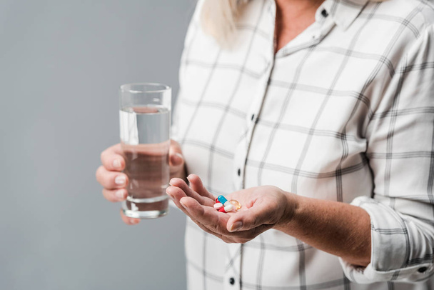 cropped view of retired woman holding pills and glass of water isolated on grey  - Foto, Imagen