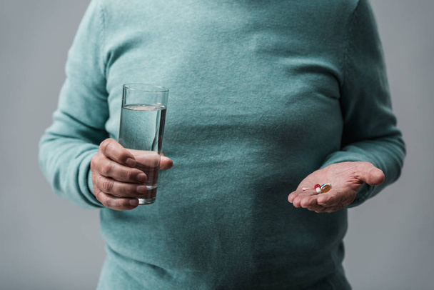 Ausgeschnittene Ansicht eines Rentners mit Tabletten und Glas Wasser isoliert auf grau  - Foto, Bild