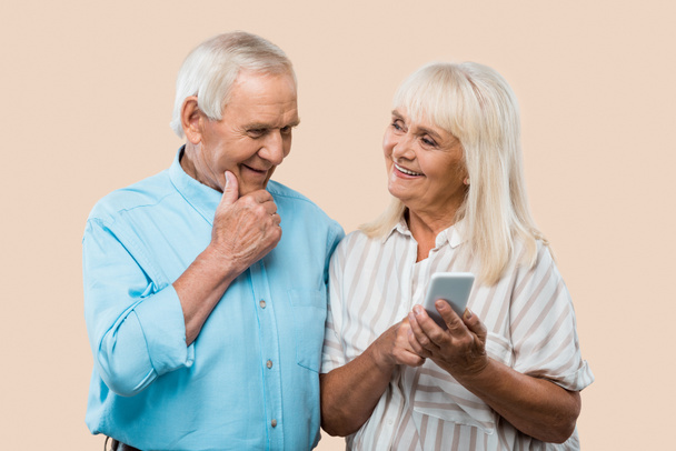 alegre jubilada sosteniendo teléfono inteligente y mirando marido aislado en beige
  - Foto, Imagen