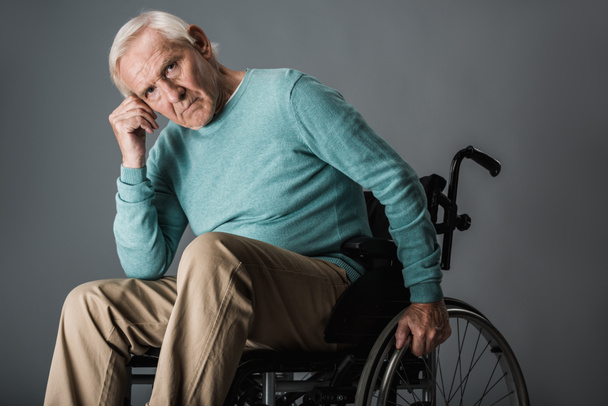 upset retired man sitting in wheelchair and looking at camera on grey - Фото, изображение