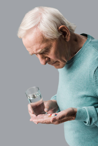 järkyttynyt eläkkeellä mies katselee pillereitä pitäen lasi vettä eristetty harmaa
  - Valokuva, kuva