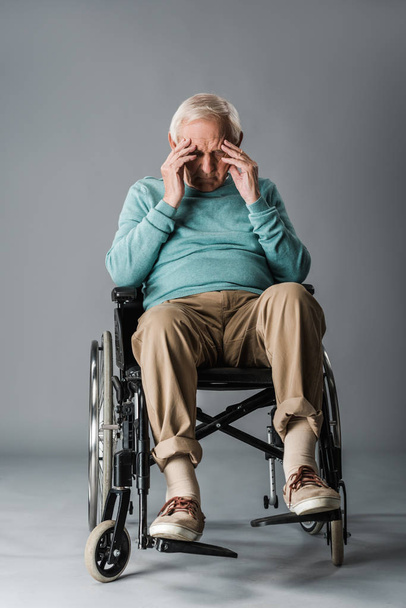 upset retired man sitting in wheelchair and touching head on grey - Fotó, kép