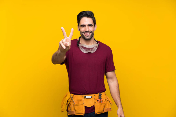schöner junger Handwerker vor isoliertem gelben Hintergrund, lächelnd und mit Siegeszeichen - Foto, Bild