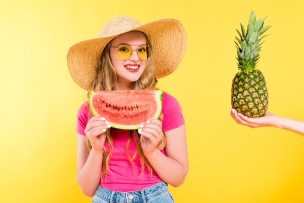 mooi meisje in stro hoed met watermeloen in de buurt vrouw vasthouden van ananas op geel - Foto, afbeelding