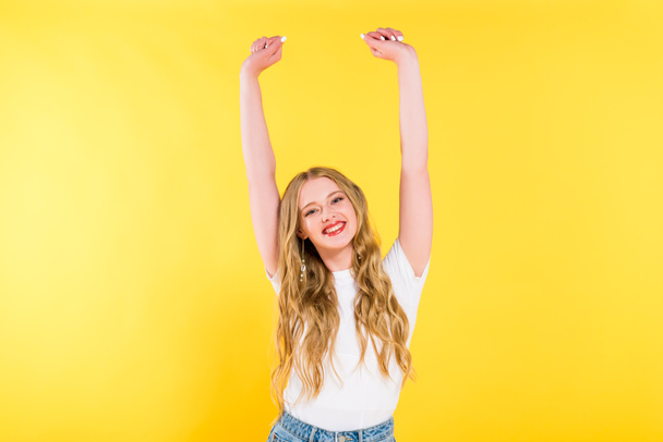 mooie gelukkige blonde jonge vrouw met verhoogde handen geïsoleerd op geel - Foto, afbeelding