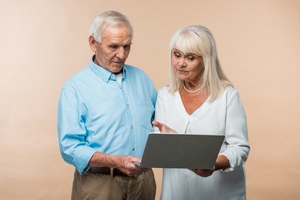 retired woman gesturing while looking at laptop near senior husband isolated on beige  - Фото, зображення