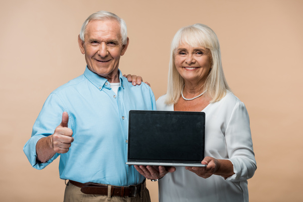 felice uomo in pensione mostrando pollice in su mentre tiene il computer portatile con schermo vuoto vicino a moglie anziana isolato su beige
  - Foto, immagini