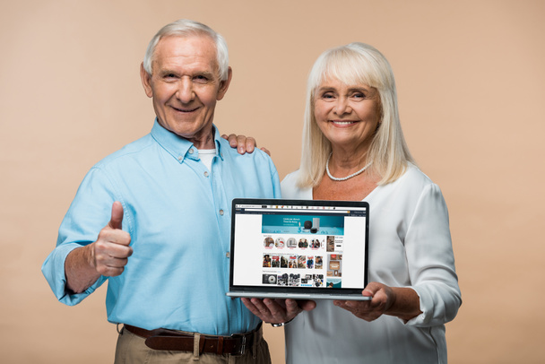 KYIV, UKRAINE - JUNE 14, 2019: happy retired man showing thumb up near wife holding laptop with amazon website on screen isolated on beige  - Foto, Bild
