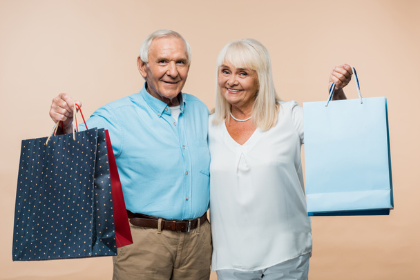 vidám nyugdíjas pár gazdaságban bevásárló táskák és mosolyogva izolált bézs  - Fotó, kép