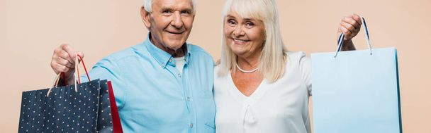 panoramic shot cheerful retired couple holding shopping bags and smiling isolated on beige  - Photo, Image