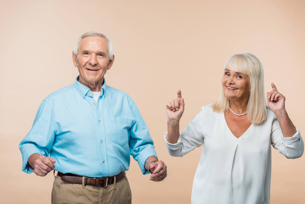 happy retired woman pointing with fingers near husband  on beige  - Foto, afbeelding