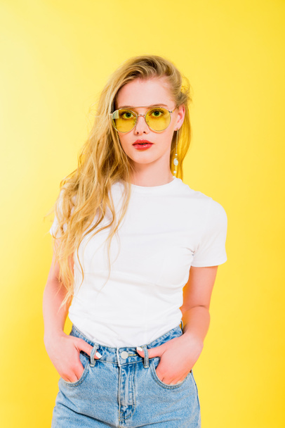 beautiful girl in sunglasses with hands in pockets looking at camera On yellow  - Photo, Image