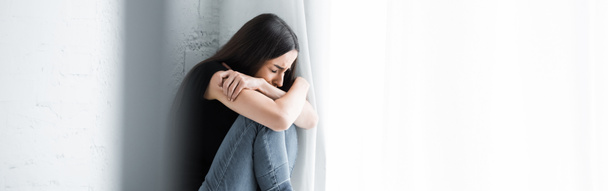 панорамний знімок пригніченої молодої жінки, що плаче, сидячи біля вікна і ховаючись у схрещених руках
 - Фото, зображення