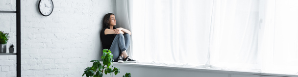 panoramische foto van depressieve jonge vrouw zittend op vensterbank met gekruiste armen en op zoek weg  - Foto, afbeelding