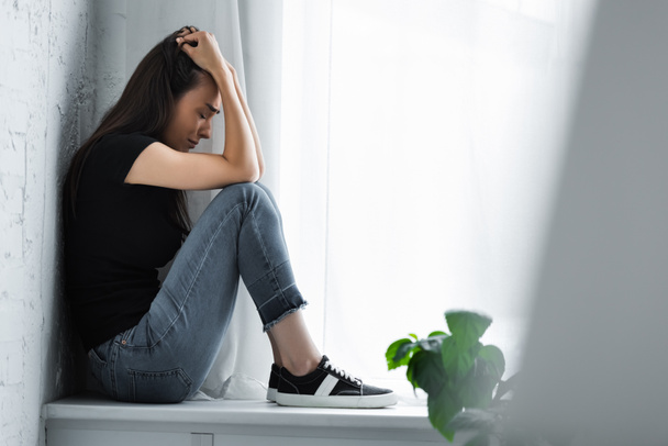 selective focus of young woman crying while sitting on window sill and suffering from depression - Foto, Bild