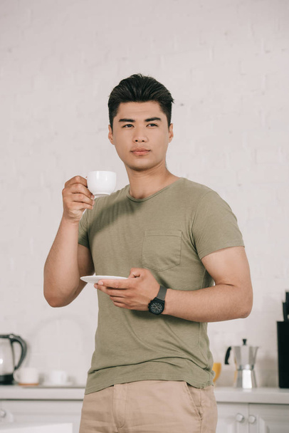 zamyślony Asian Mężczyzna picie kawy stojąc w kuchni i patrząc z dala - Zdjęcie, obraz