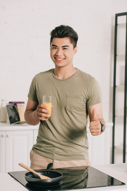 sonriente asiático hombre celebración de vaso de naranja jugo y mostrando el pulgar hacia arriba mientras sonriendo a la cámara
 - Foto, Imagen