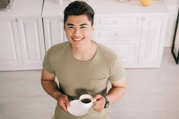 overhead view of cheerful asian man holding coffee cup while smiling at camera - Foto, afbeelding