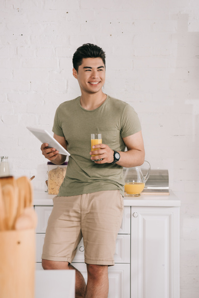 красивый азиатский мужчина держит стекло с апельсиновым соком и цифровой планшет, улыбаясь и глядя в сторону на кухне
 - Фото, изображение