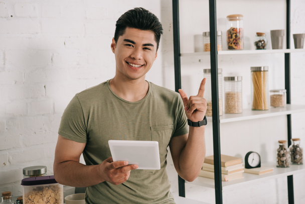 cheerful asian man holding digital tablet and showing idea sign while smiling at camera - Zdjęcie, obraz