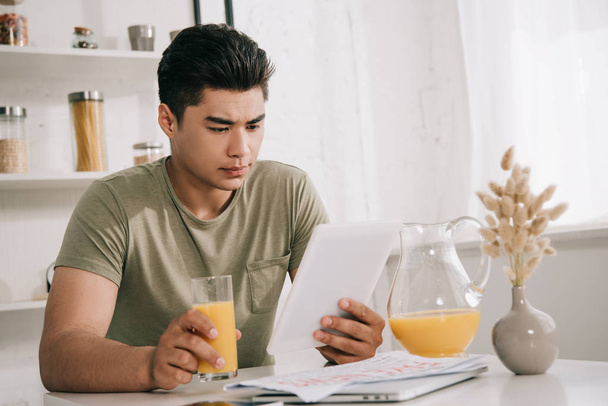 handsome asian man looking at digital tablet while sitting at kitchen table and holding glass of orange juice - Photo, Image