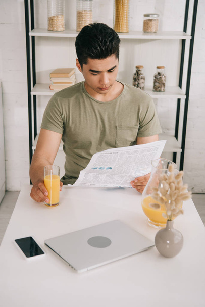 tarkkaavainen aasialainen mies lukee sanomalehteä istuessaan keittiön pöydässä ja pitäen lasillista appelsiinimehua
 - Valokuva, kuva