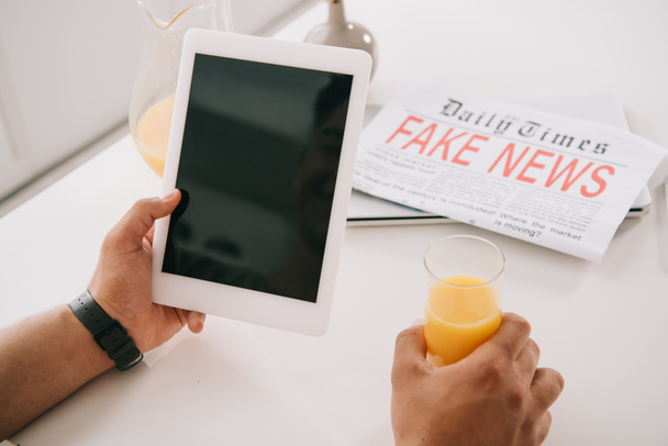 cropped view of man holding glass of orange juice and digital tablet with blank screen while sitting at kitchen table near fake news newspaper - Foto, Bild