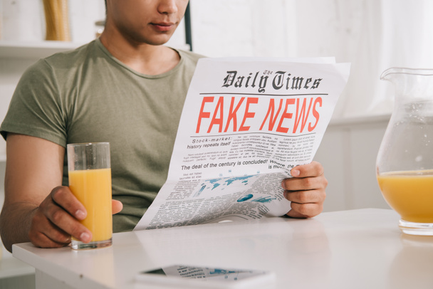 cropped view of young man reading fake news newspaper while sitting at kitchen table and holding glass of orange juice - 写真・画像